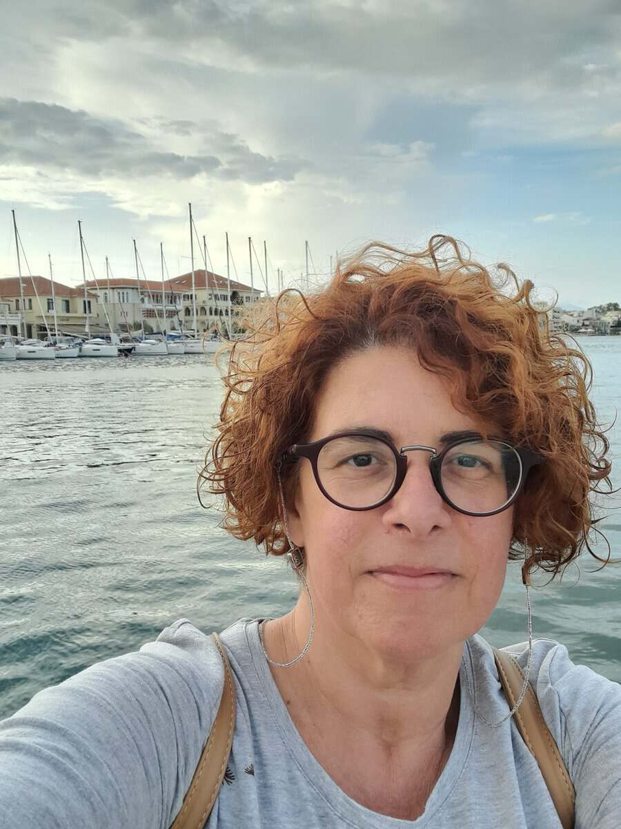 Γυναίκα με καστανά σγουρά μαλλιά και γυαλιά, μπροστά από λιμάνι με ιστιοπλοϊκά σκάφη και ουρανό με σ.